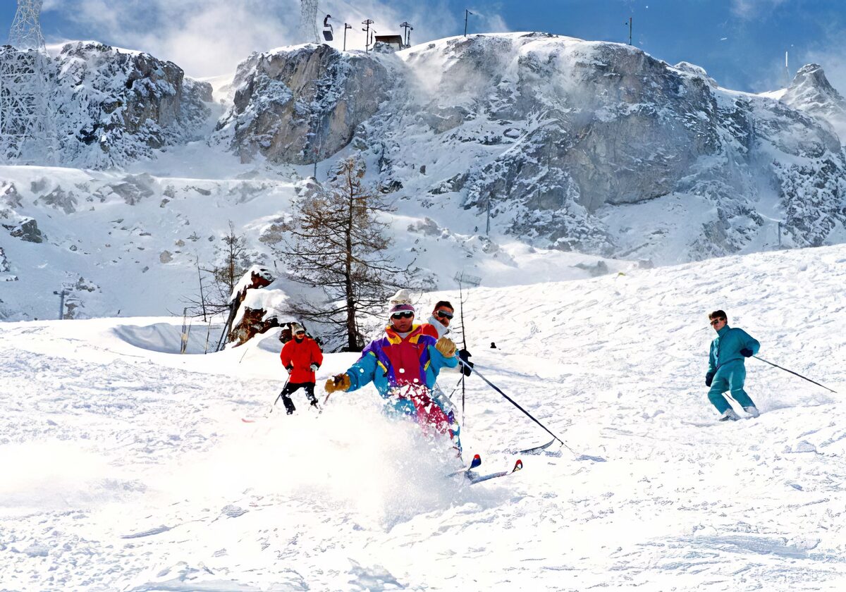 Skiers in powder on a steep snow slope below a rocky ridge, with a lift station at the top