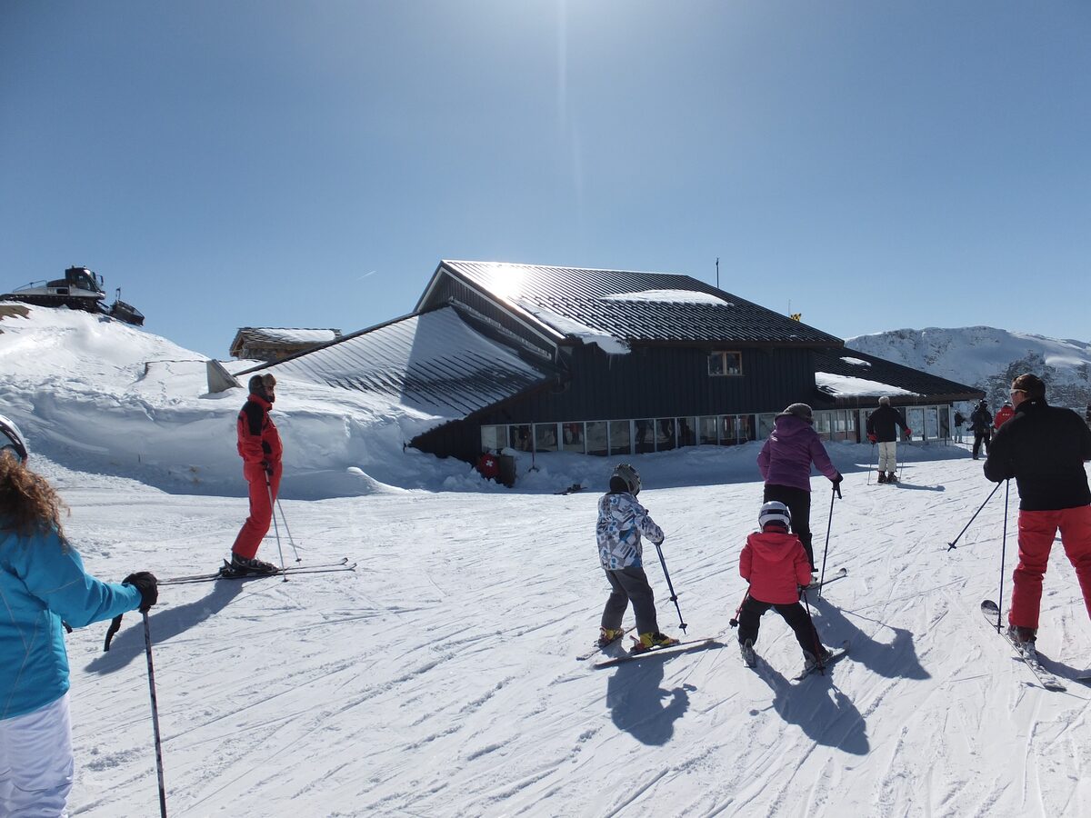 Skiers set off across a sunny snowy slope next to the mountain restaurant at the top of the Funival