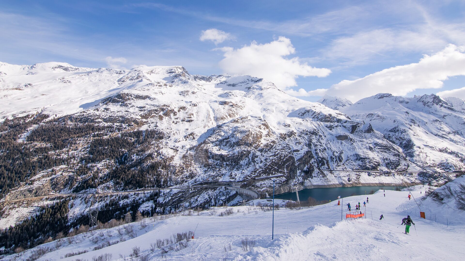 Skiers on a snowy piste above the Chevril lake and the Tignes dam, with snow-covered mountains behind