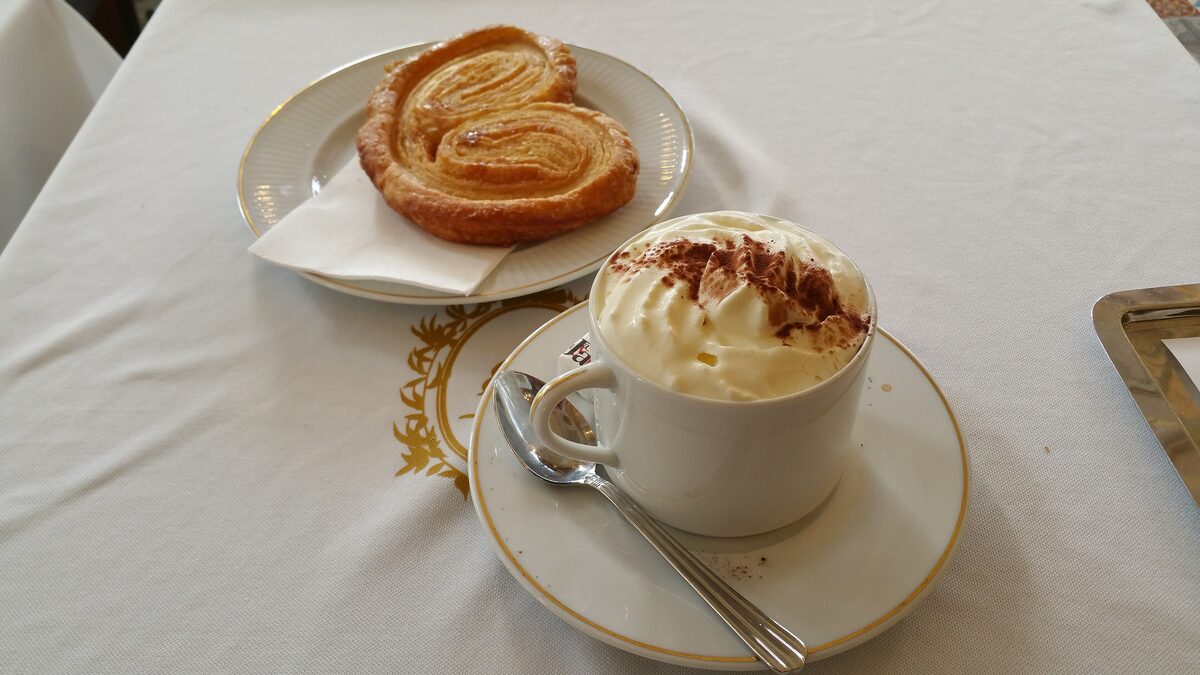 A cup of hot chocolate with whipped cream and cocoa next to a palmier pastry
