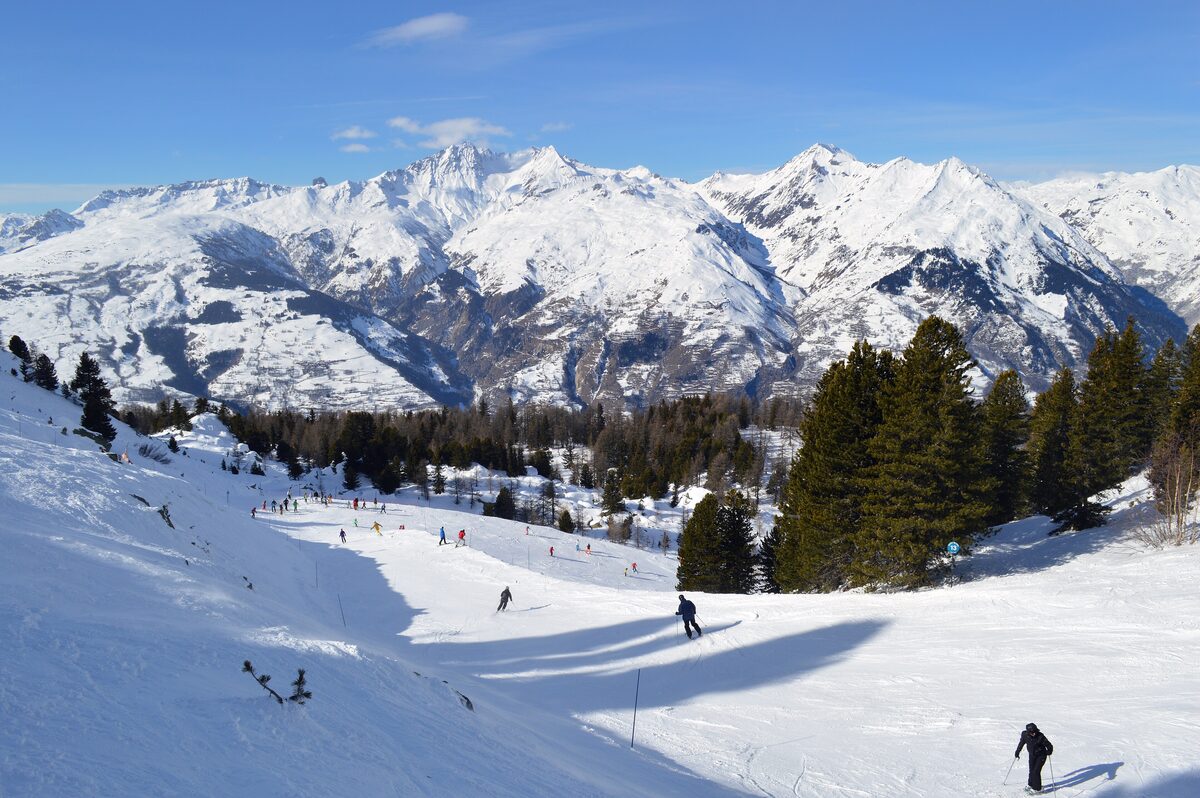 Skiers on a sunny, snow-covered slope at Les Arcs with snowy peaks behind