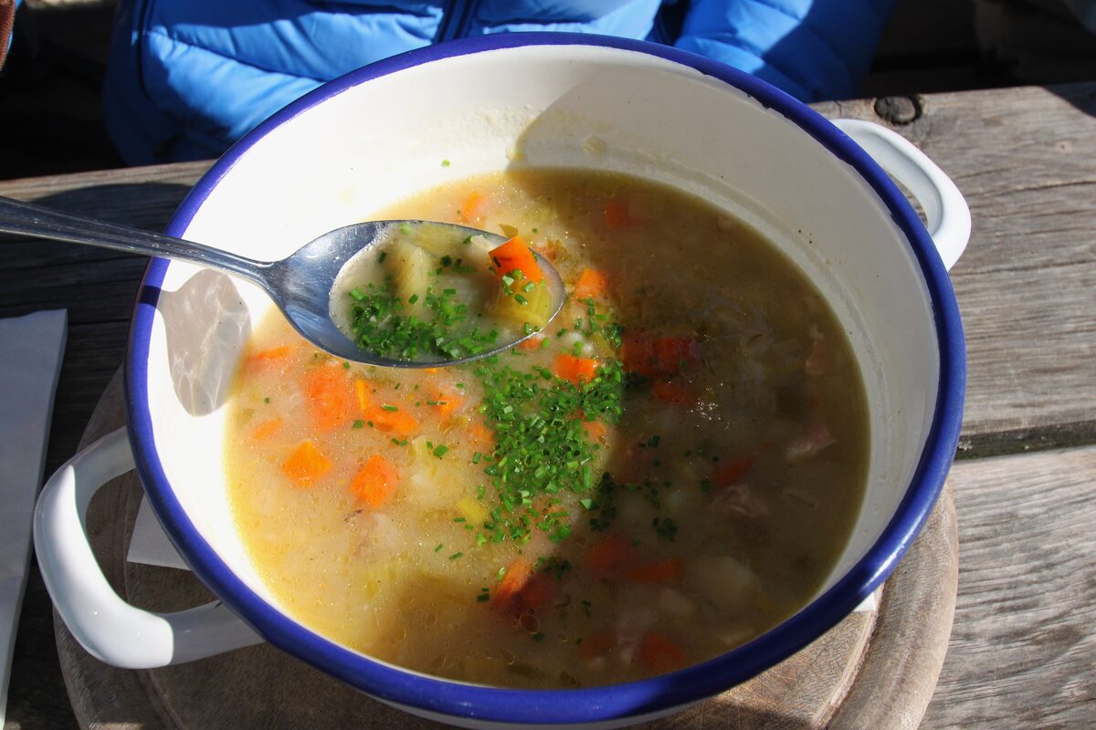 A pot of barley soup with carrots, potatoes and parsley on a wooden hut table, a spoon in the bowl