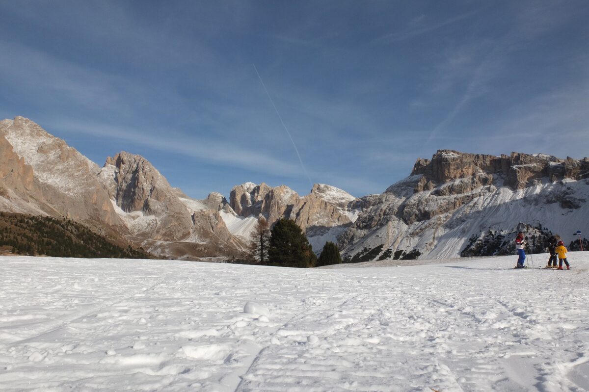 A fresh snowfield on the slopes above Santa Cristina with two skiers at the edge and the rock walls of the Odle group rising behind; the village itself is below the frame