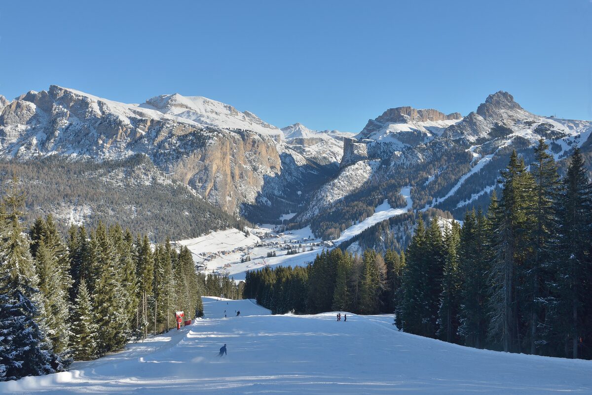 The Ciampinoi piste running down toward Selva in the valley, with snowy Dolomite peaks across the valley under a blue sky