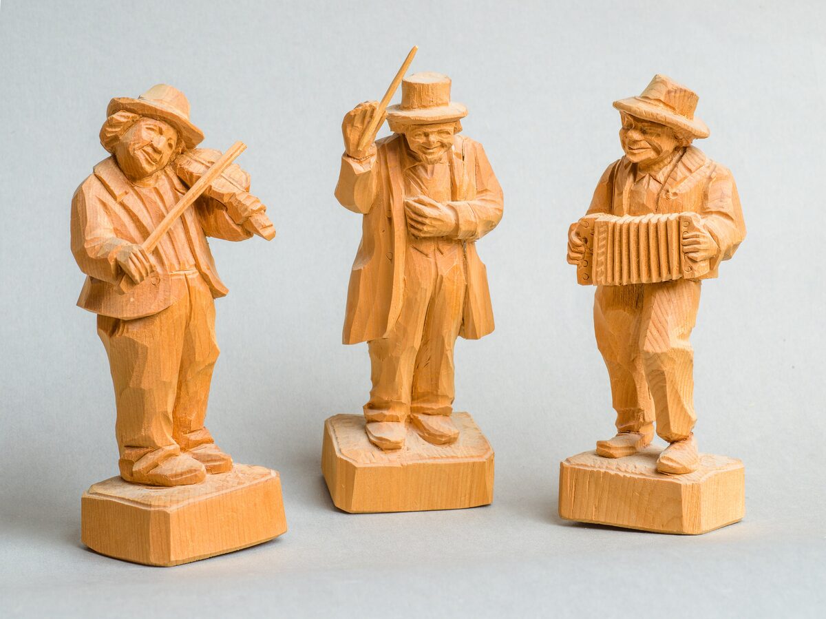Three small hand-carved wooden figures from Val Gardena, a fiddler, a conductor and an accordion player, in pale pine on a plain background
