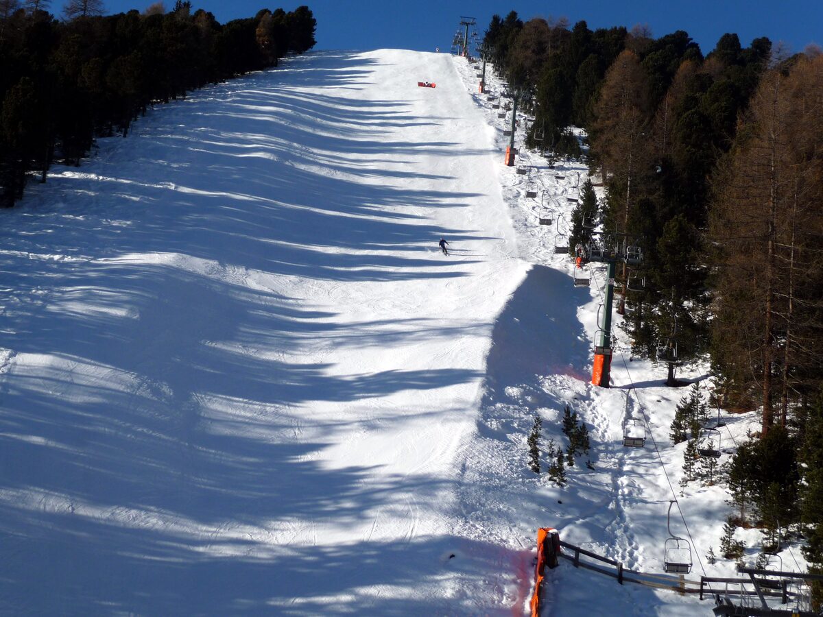 A black piste below the Col Raiser refuge in fresh snow, with trees lining the run and a skier on the slope