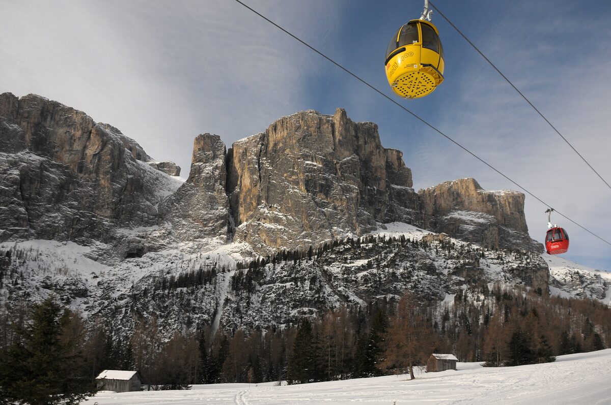 A yellow gondola cabin and a red one on the cable above snowy slopes, with the rock walls of the Sella group behind, on the way from Passo Gardena toward Colfosco