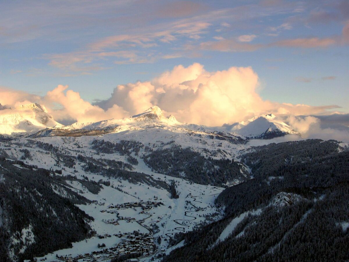 The snow-filled Corvara valley in Alta Badia seen from above in late light, with mountains and clouds glowing beyond