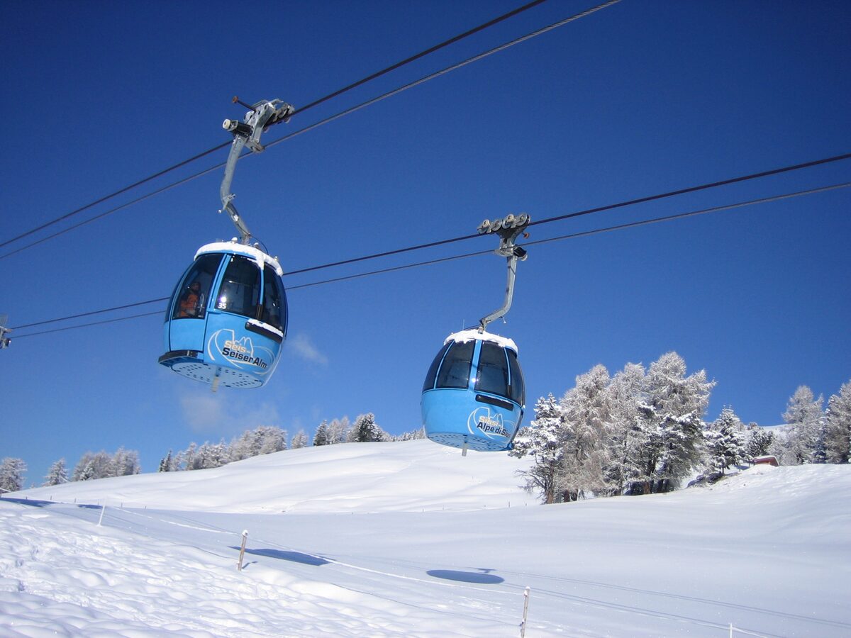 Two blue Alpe di Siusi gondola cabins on the cable above a snow-covered pasture and frosted larches, under a deep blue sky