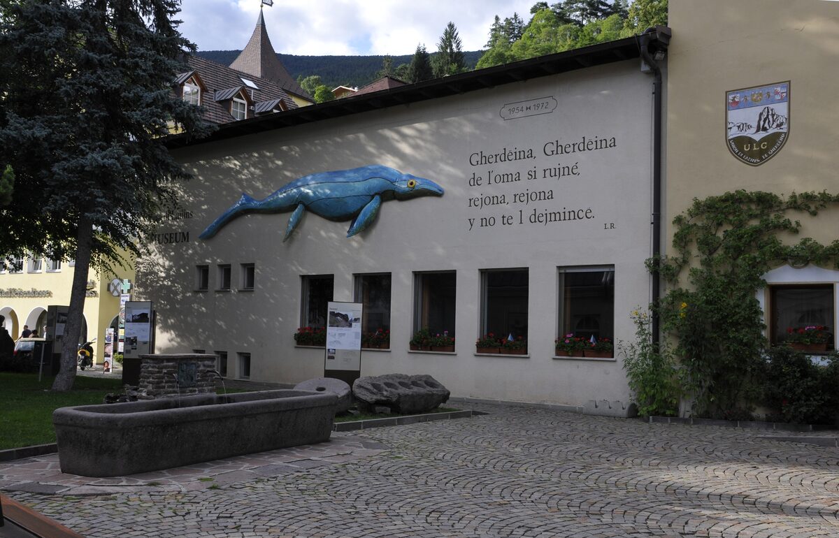 The Museum Gherdëina building in Ortisei, with a blue sculpture of a marine reptile and a Ladin slogan on the wall