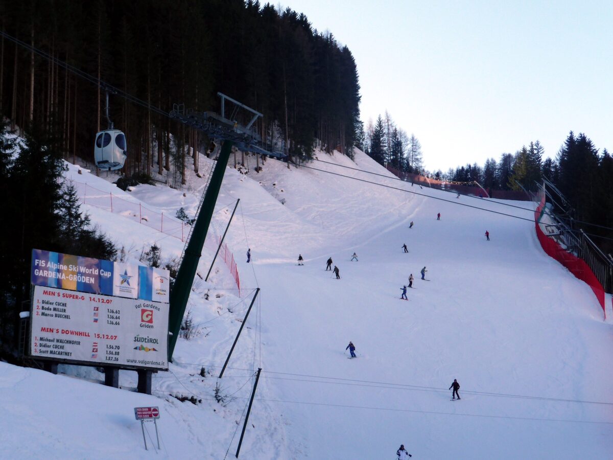 Skiers on the wide Saslong piste in winter, with race fencing and a timing board beside the lift line