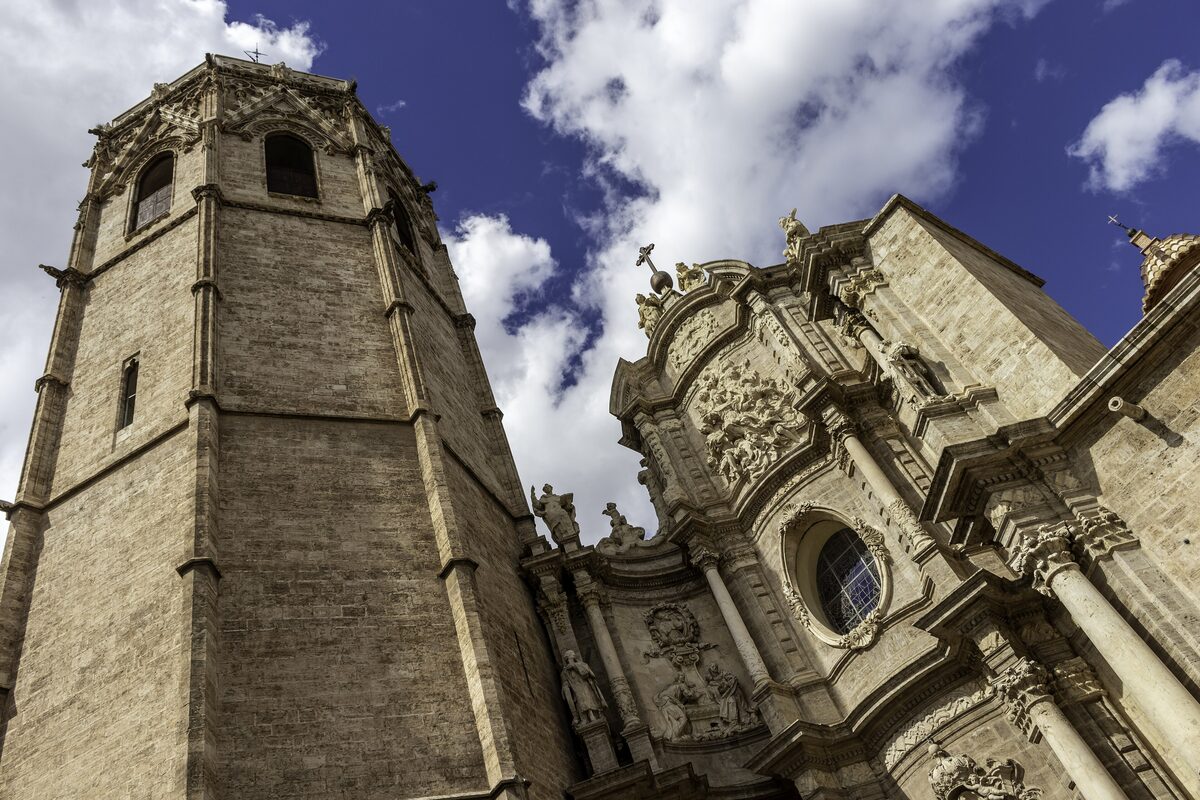 The octagonal Gothic bell tower of the Micalet next to the baroque facade of Valencia Cathedral, under blue sky with white clouds