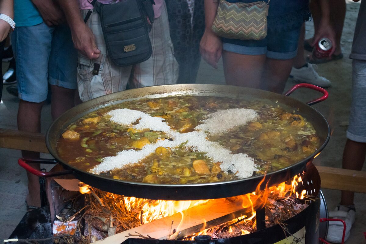 A large paella pan cooking over a wood fire, with rice being added to the stock in the shape of a cross