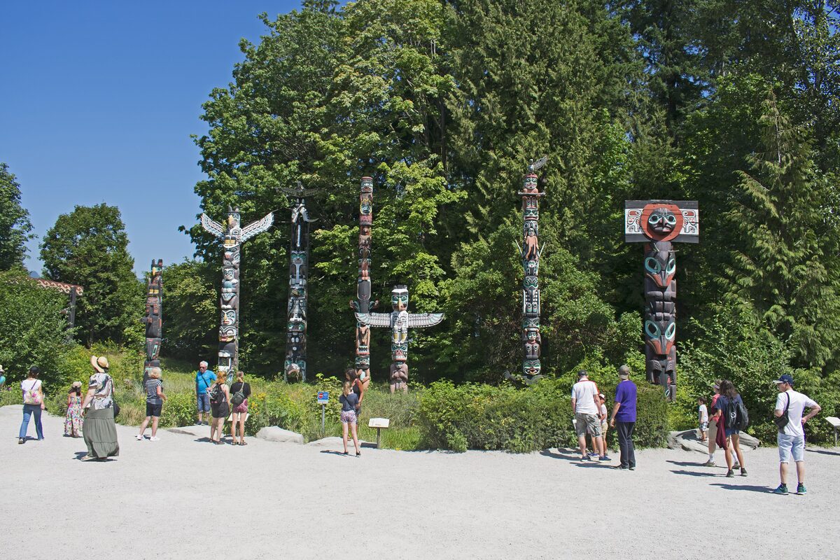 Five carved totem poles standing in a row on a gravel plaza at Brockton Point, with visitors in front and tall trees behind