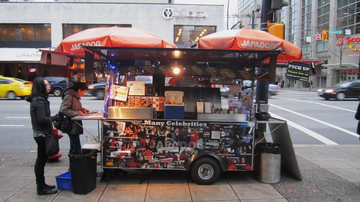 A Japadog street cart with orange umbrellas on a downtown Vancouver corner, with customers at the counter
