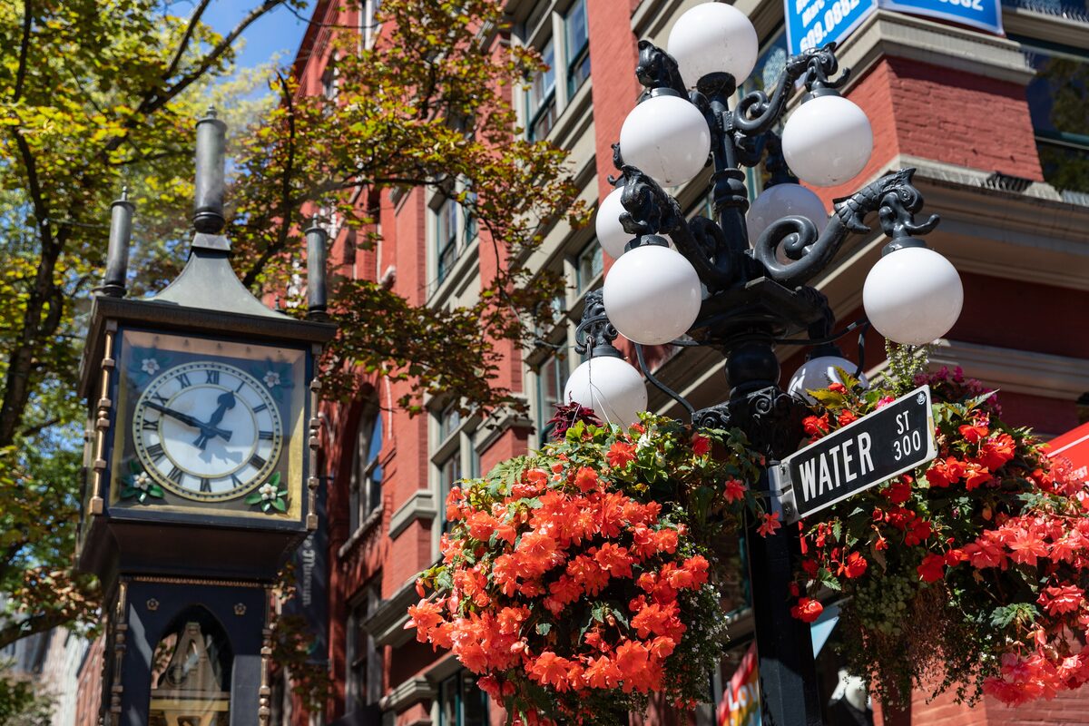 The Gastown steam clock on Water Street beside a lamp post hung with red flower baskets and brick buildings