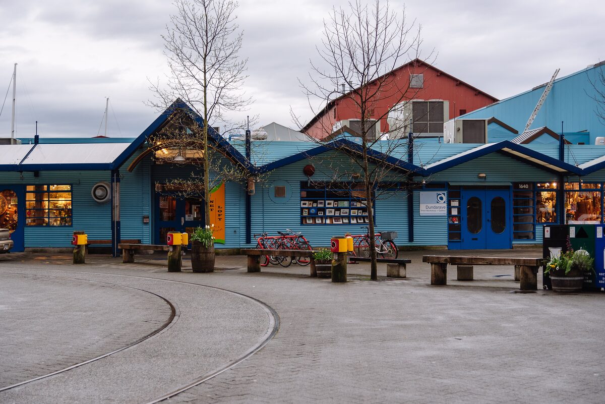 Blue and red industrial sheds on Granville Island, now studios and shops, with old rail tracks in the paving