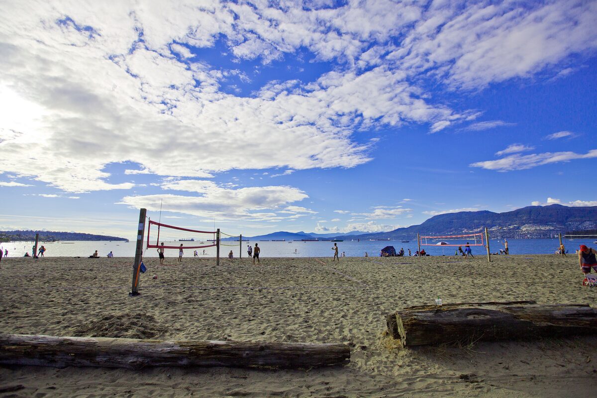 Kitsilano Beach with a volleyball net and driftwood logs on the sand, and the North Shore mountains across the bay