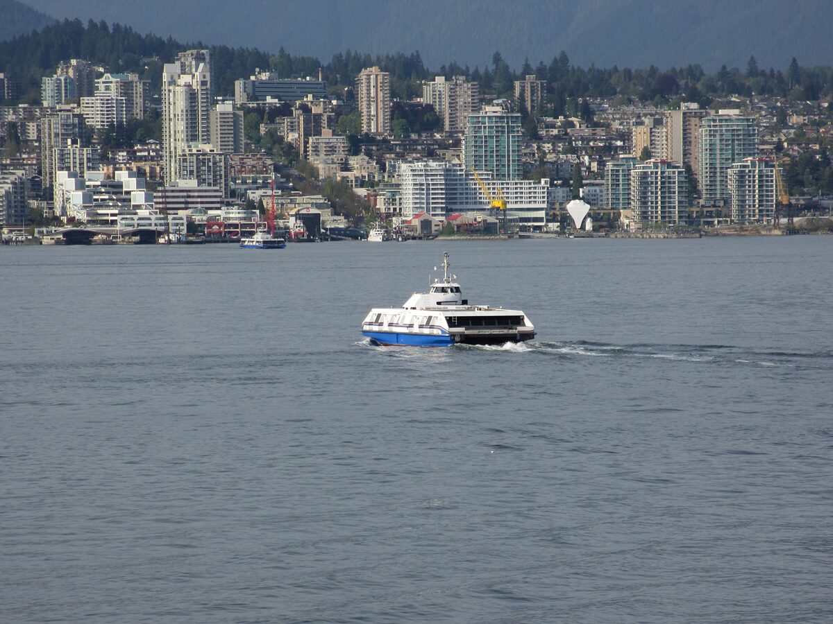 A SeaBus catamaran ferry crossing Burrard Inlet with the North Vancouver waterfront and forested hills behind