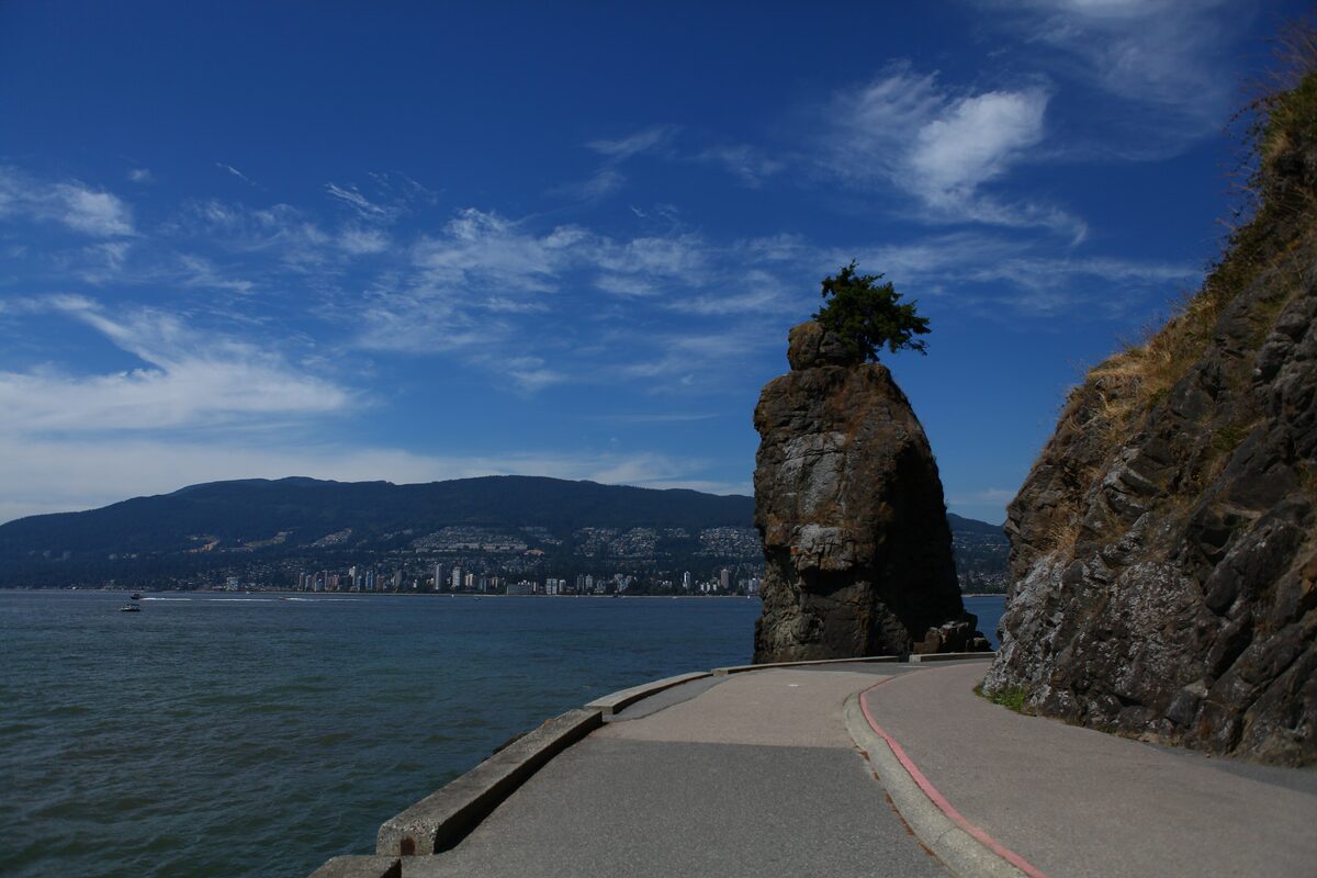 Siwash Rock, a rock pillar topped with a small tree, beside the Seawall path with the North Shore mountains across the water