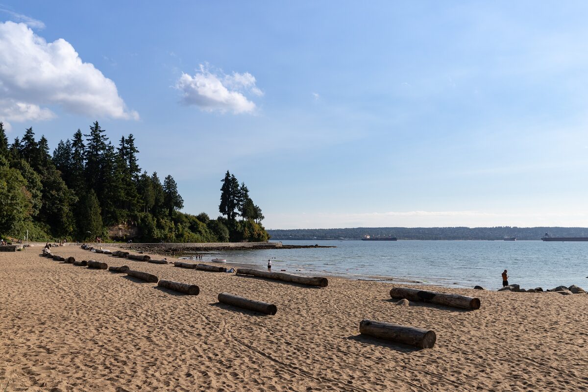 Third Beach in Stanley Park, a wide sand beach scattered with driftwood logs, with forest on the point and the strait beyond
