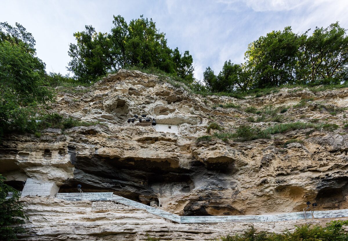 The limestone cliff of the Aladzha rock monastery with carved cells, caves and walkways, topped by forest.