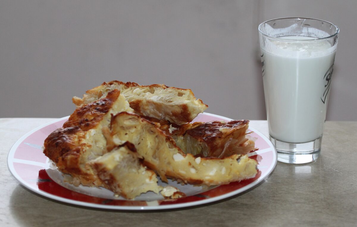 Flaky pieces of golden cheese banitsa on a plate next to a tall glass of white yogurt drink.