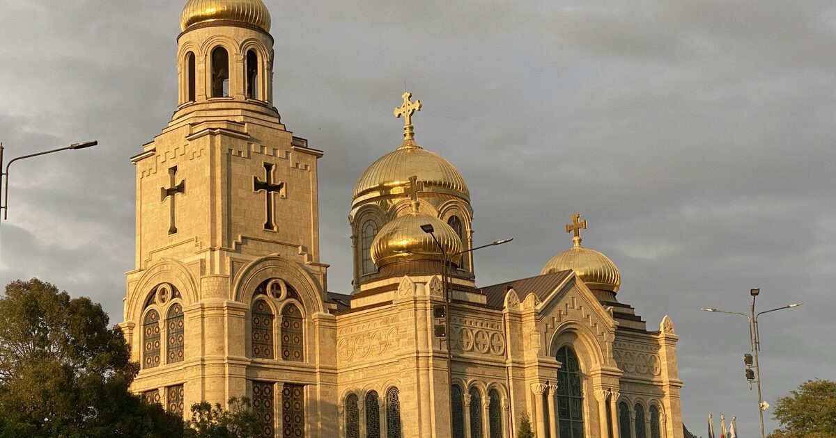 The Dormition Cathedral in Varna with its gilded domes lit by golden evening sun against a grey sky.