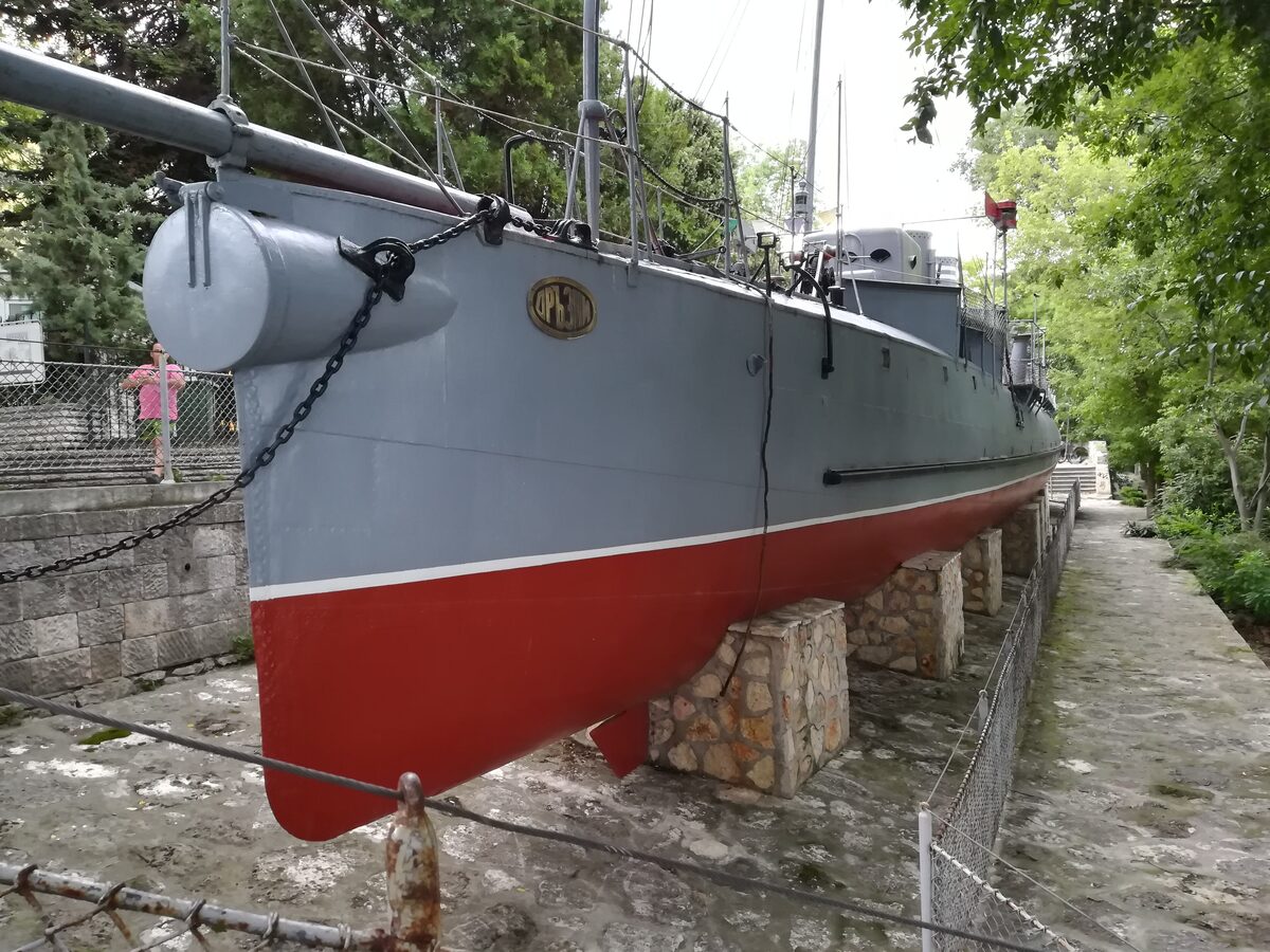 The grey and red hull of the torpedo boat Drazki, its name plate on the bow, resting on stone supports in the Naval Museum yard.
