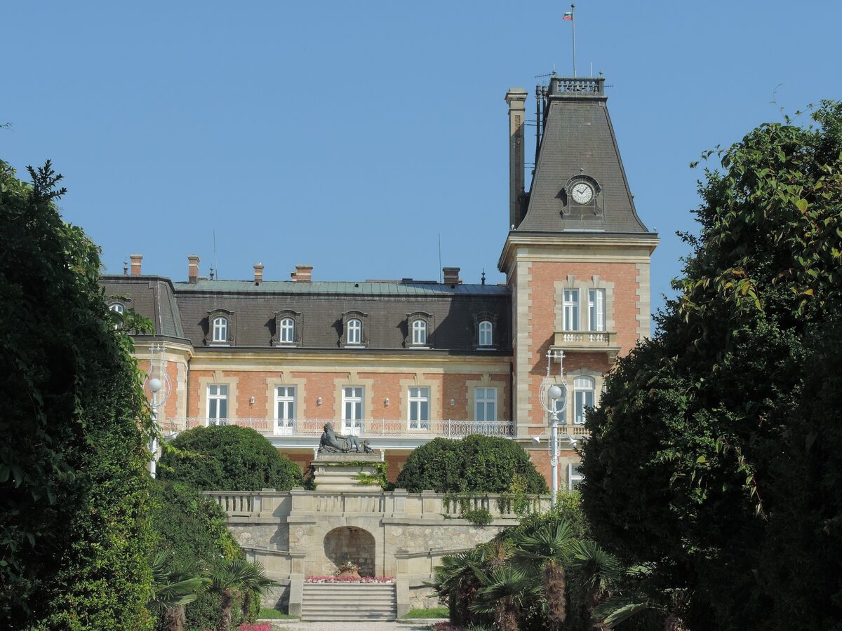The brick-and-slate Euxinograd palace with its clock tower, seen up a garden alley past the Neptune statue and terrace steps.
