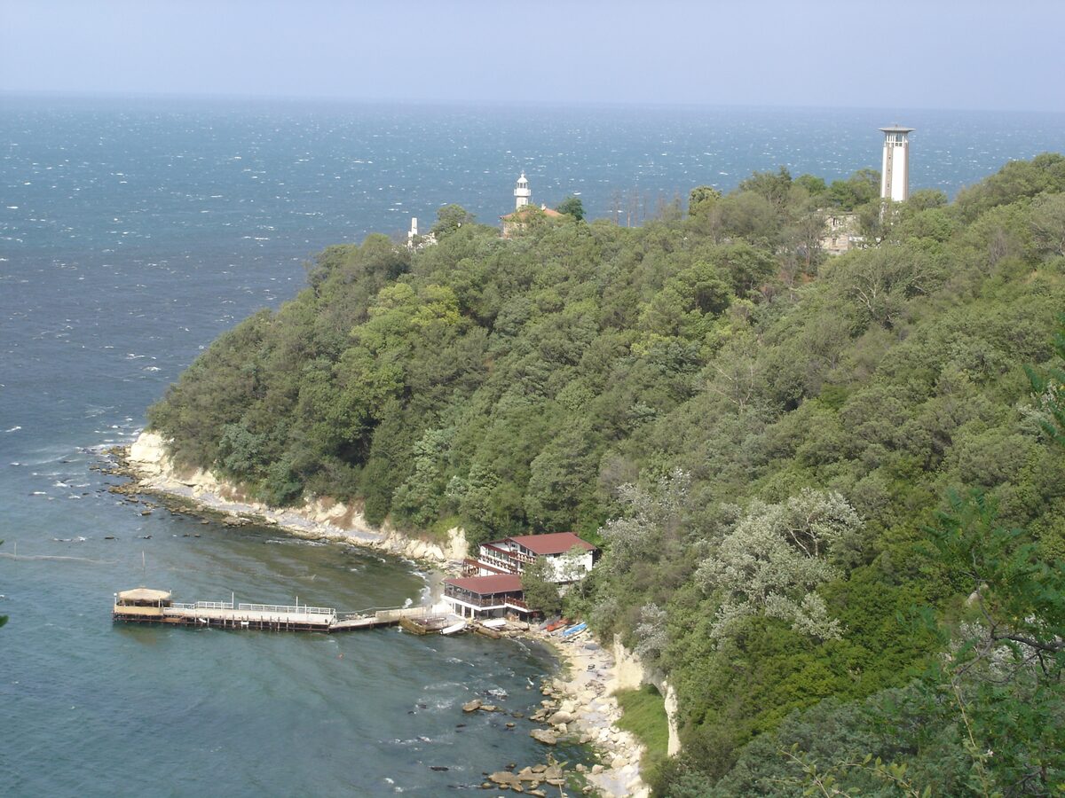 Wooded Cape Galata dropping to the sea, with lighthouses on top and a small pier and restaurant at the foot of the cliffs.