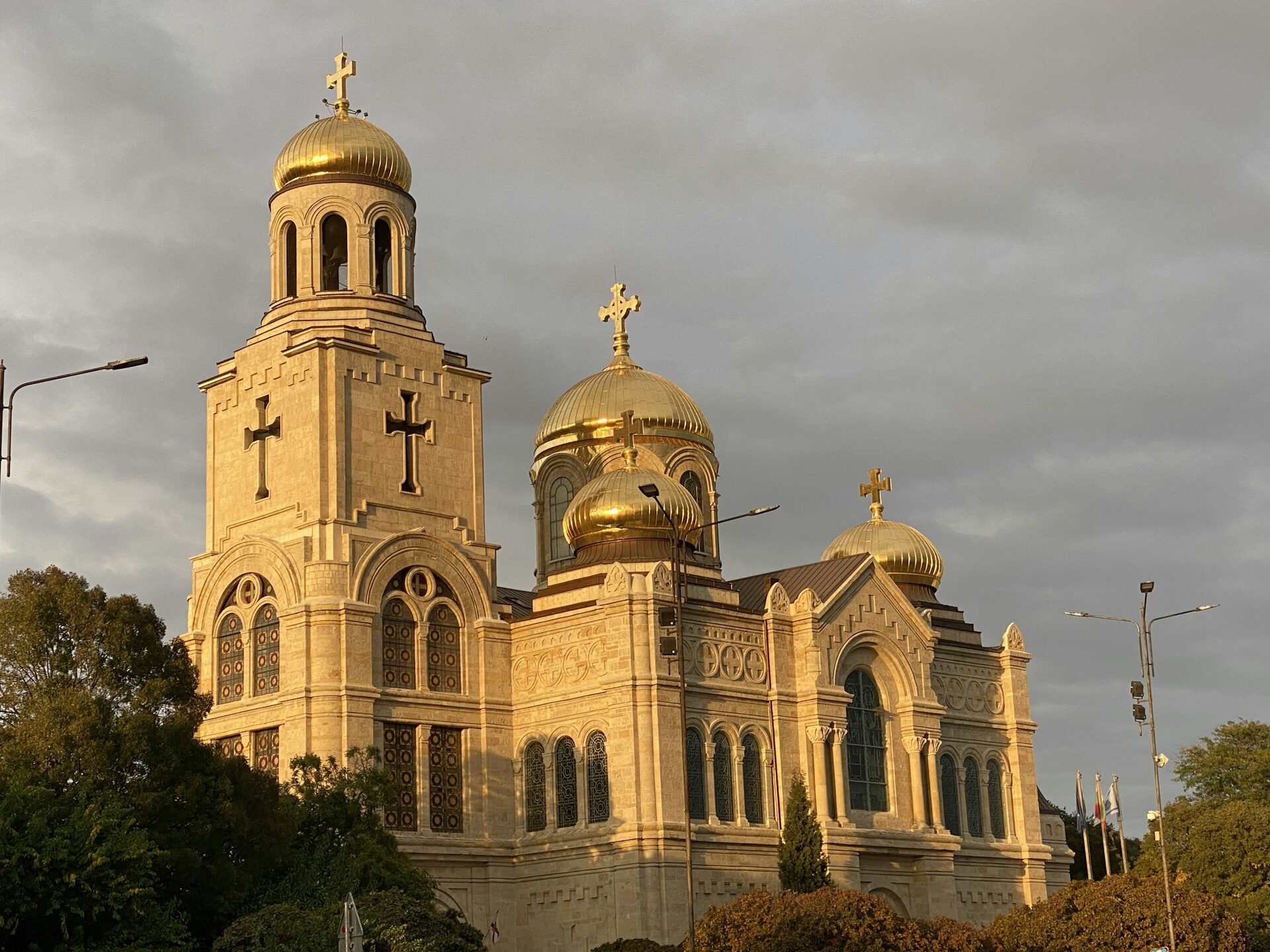 The Dormition Cathedral in Varna with its gilded domes lit by golden evening sun against a grey sky.