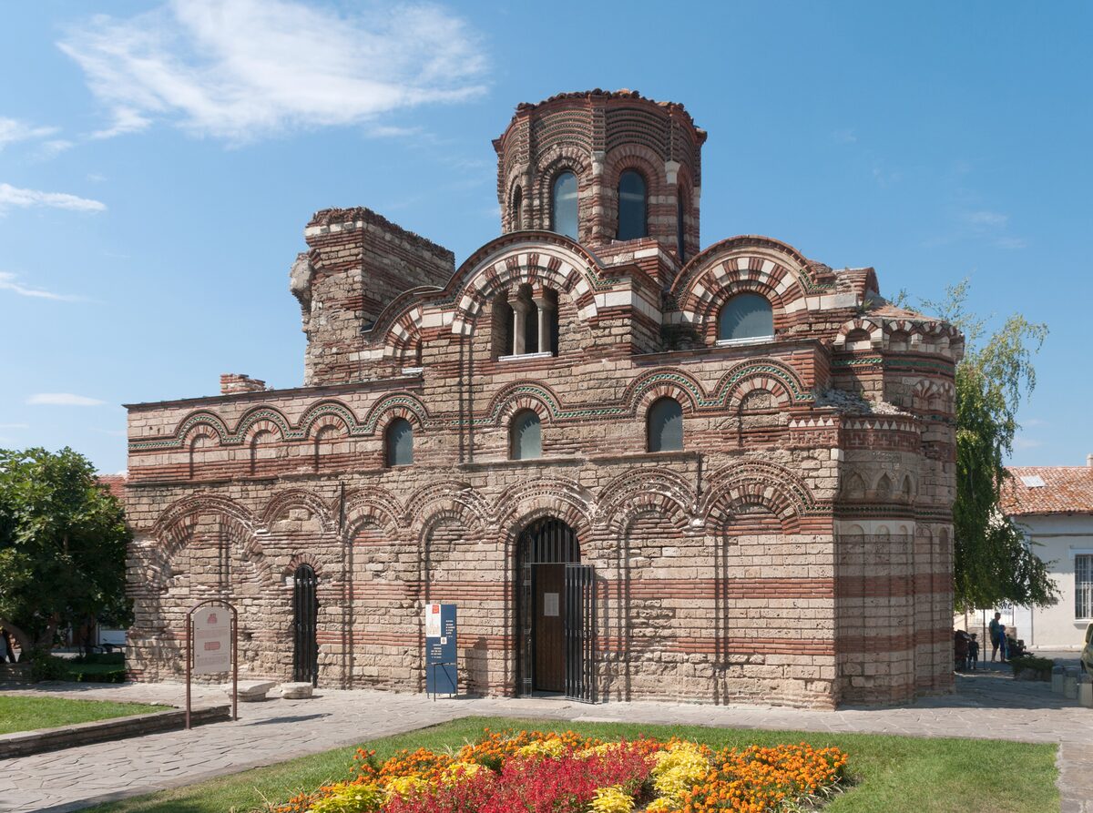 The 13th-14th century Church of Christ Pantocrator in Nesebar, its banded brick and stone walls decorated with blind arches, behind a flowerbed.