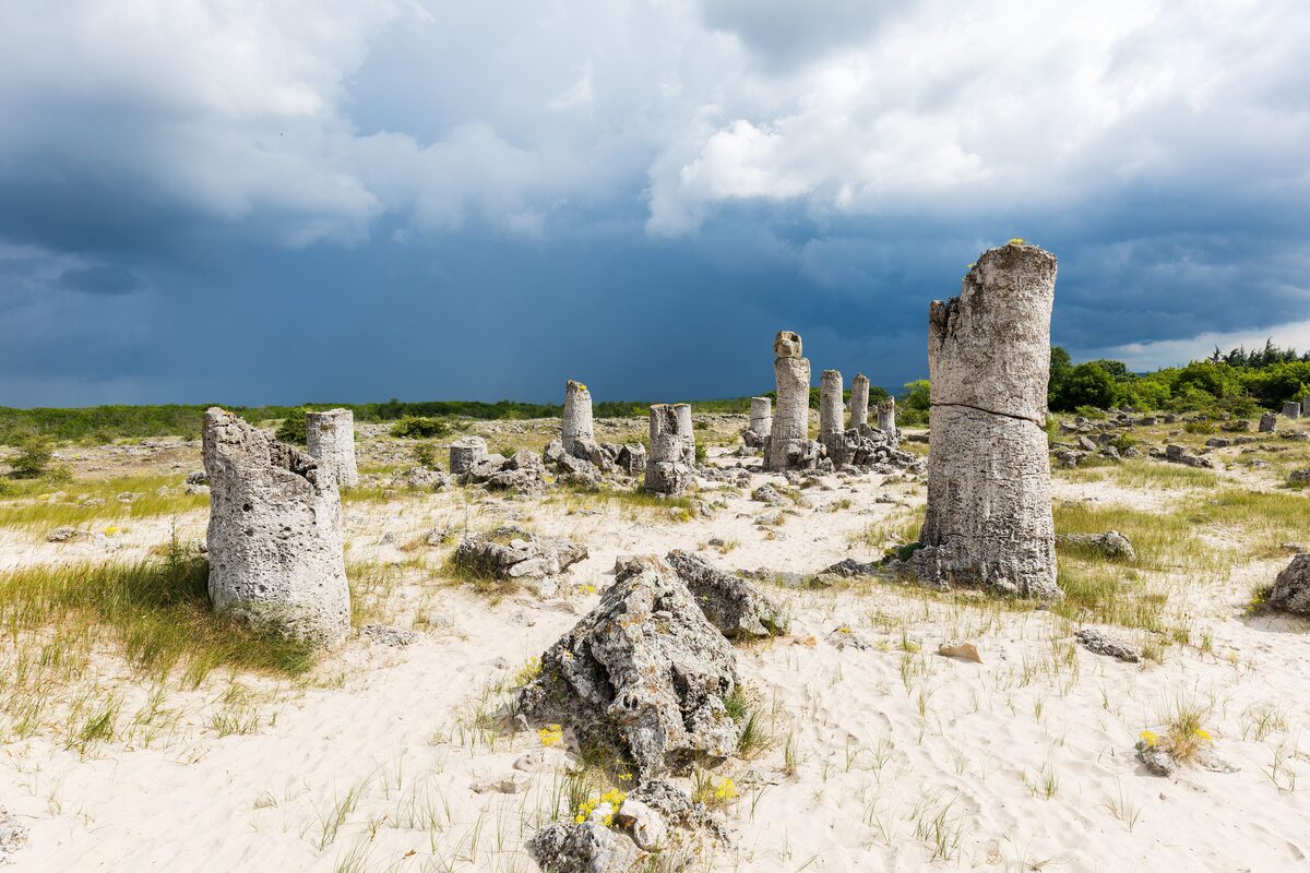 Stone columns of Pobiti Kamani rising from white sand and grass under a dark storm sky.