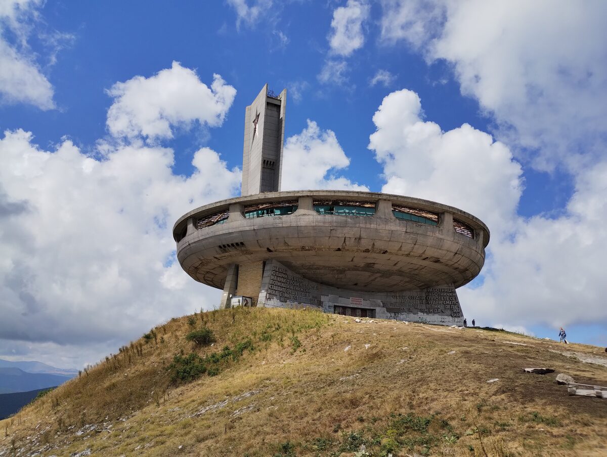 The saucer-shaped concrete Buzludzha monument and its tall tower with a red star, on a grassy peak under a cloudy blue sky.