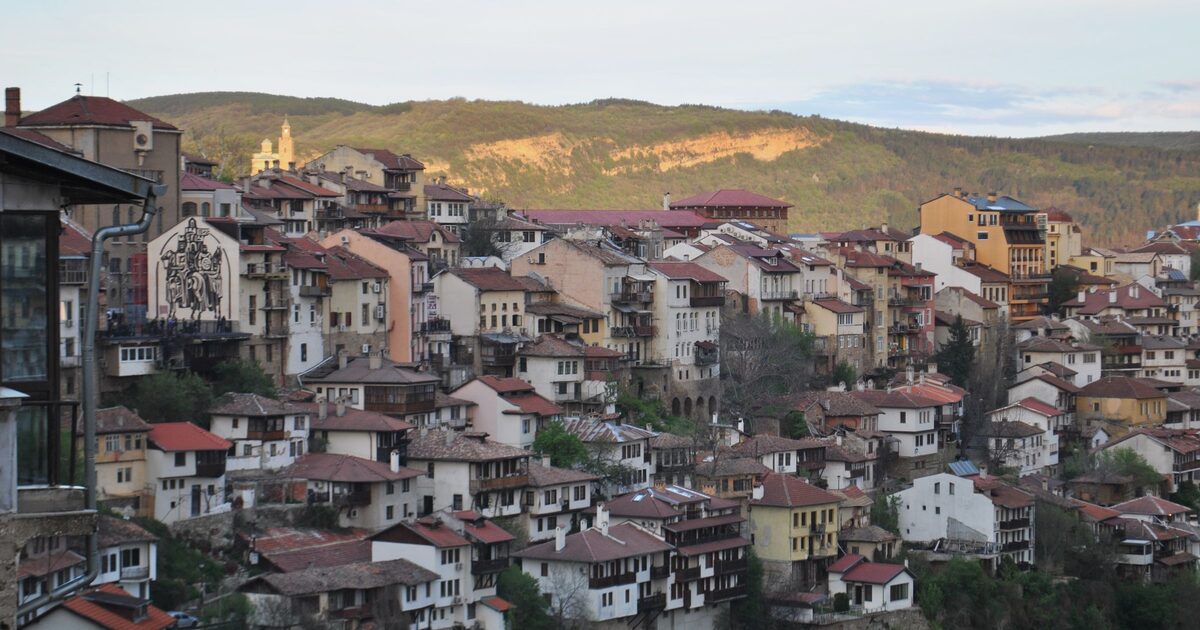 Old houses of Veliko Tarnovo stacked on the cliffs above the Yantra gorge, with green hills behind in late-afternoon light.
