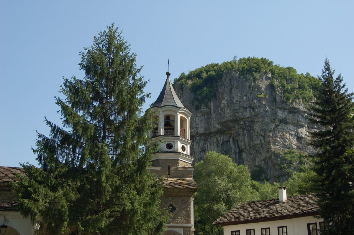 The bell tower of Dryanovo Monastery between tall firs, with a sheer limestone cliff of the Andaka gorge rising behind.