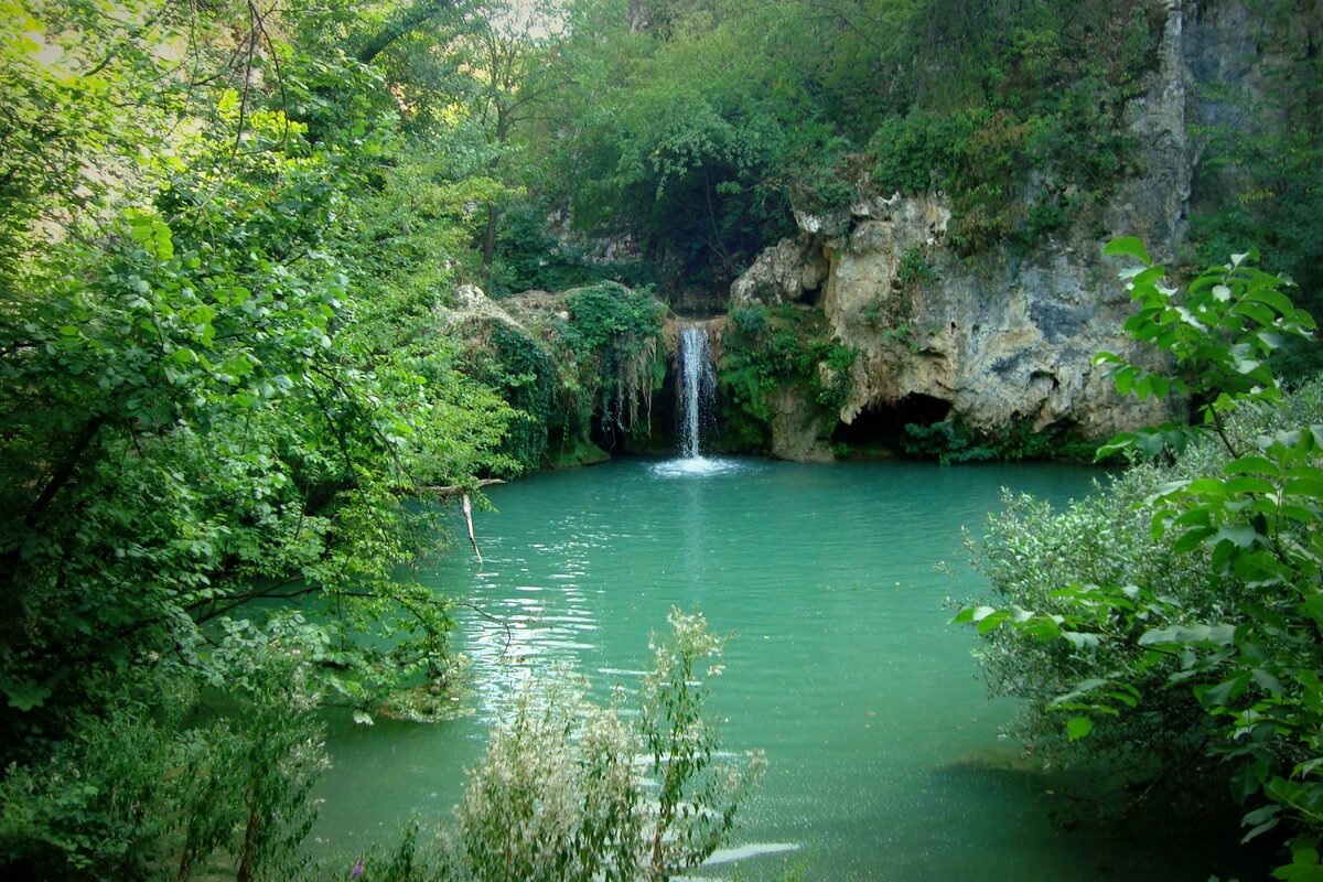 The Hotnitsa waterfall dropping from a limestone ledge into a turquoise pool, framed by dense green foliage.