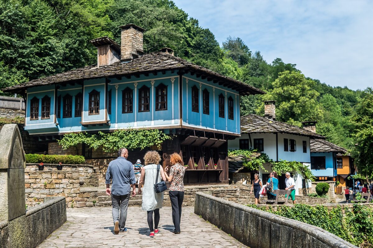 Visitors crossing a stone bridge towards a sky-blue Revival-era house with a stone-slab roof at the Etar open-air museum, with wooded hills behind.