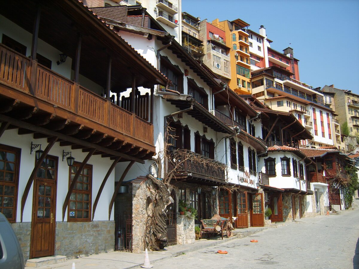 Whitewashed Revival-era houses with carved wooden balconies and eaves along cobbled Gurko street, with taller buildings stepping up the hill behind.