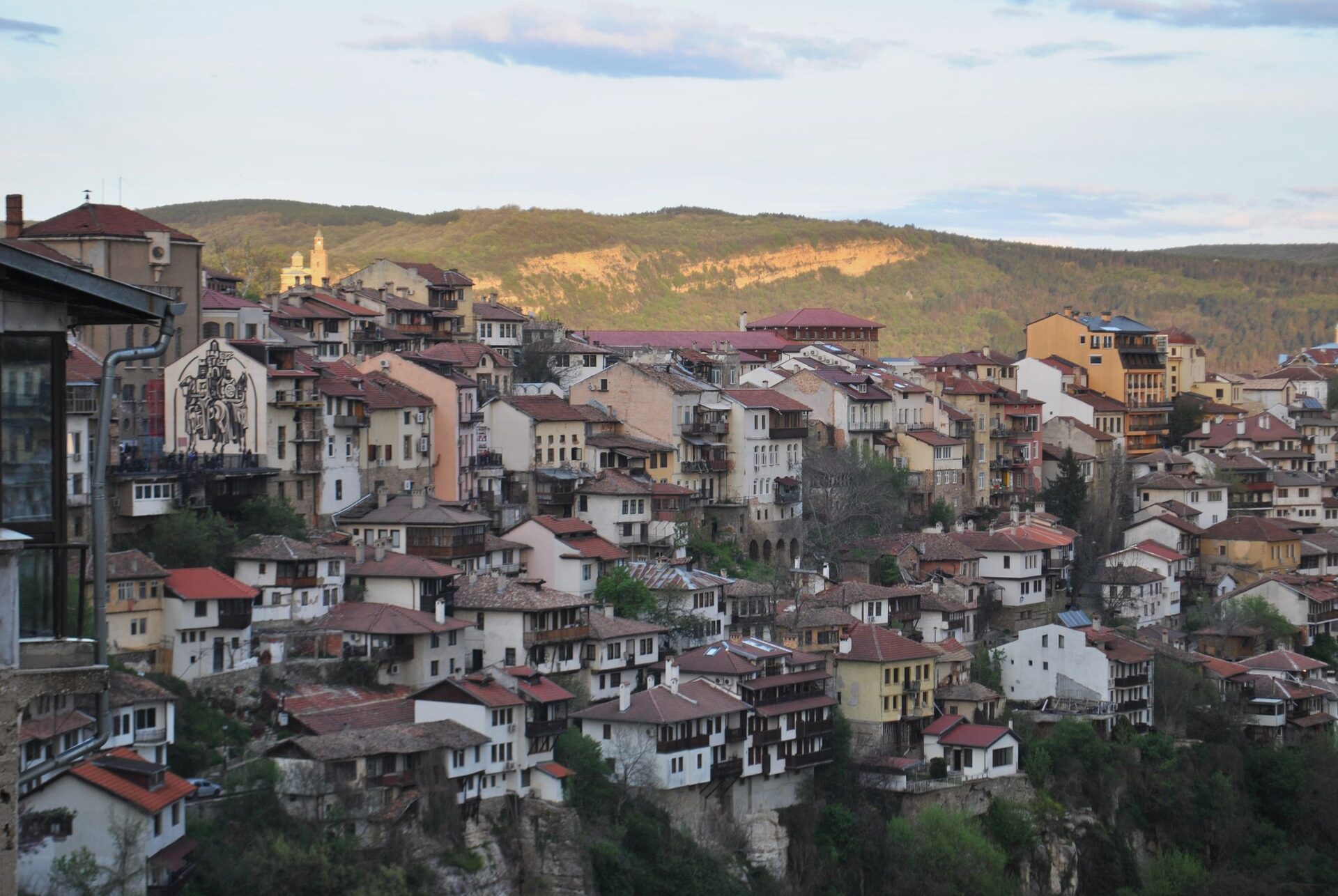 Old houses of Veliko Tarnovo stacked on the cliffs above the Yantra gorge, with green hills behind in late-afternoon light.