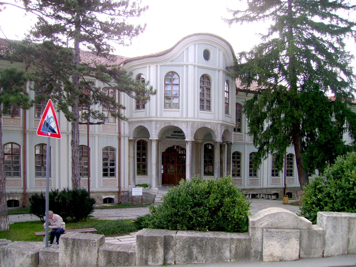 The pale Konak building, now the Museum of the Revival and Constituent Assembly, with its curved central bay and arcaded entrance framed by tall conifers.