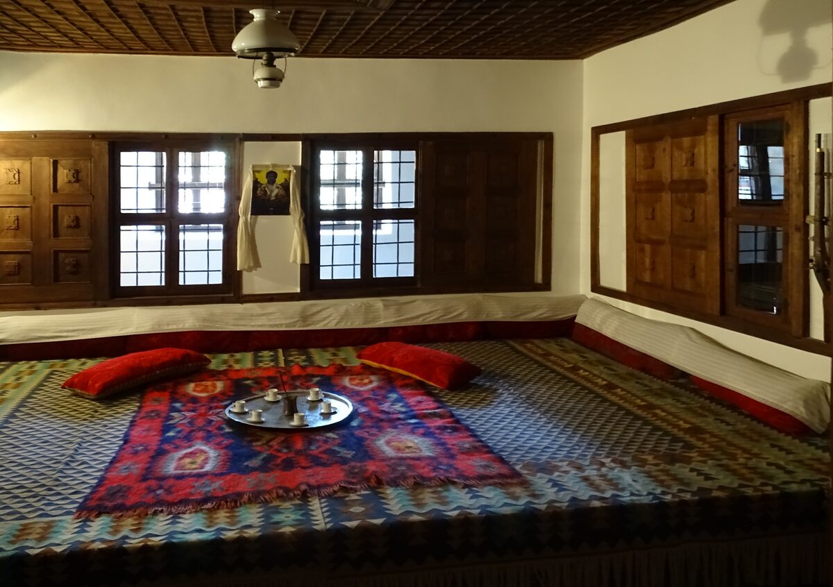 A furnished room in the Konstantsalieva house in Arbanasi, with a low cushioned platform under woven kilims, a coffee tray, carved wooden shutters and a coffered wooden ceiling.