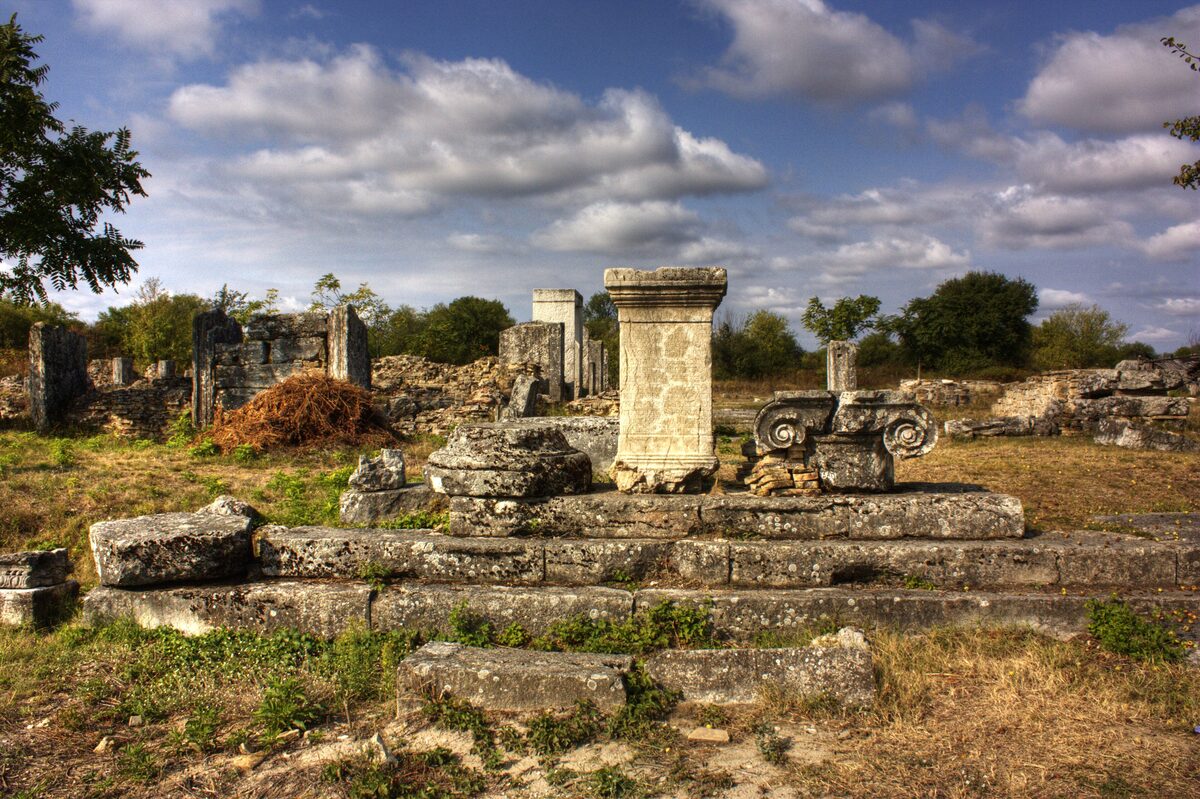 Stone steps, a carved pedestal and an Ionic capital among the ruins of the Roman town of Nicopolis ad Istrum under a cloudy blue sky.