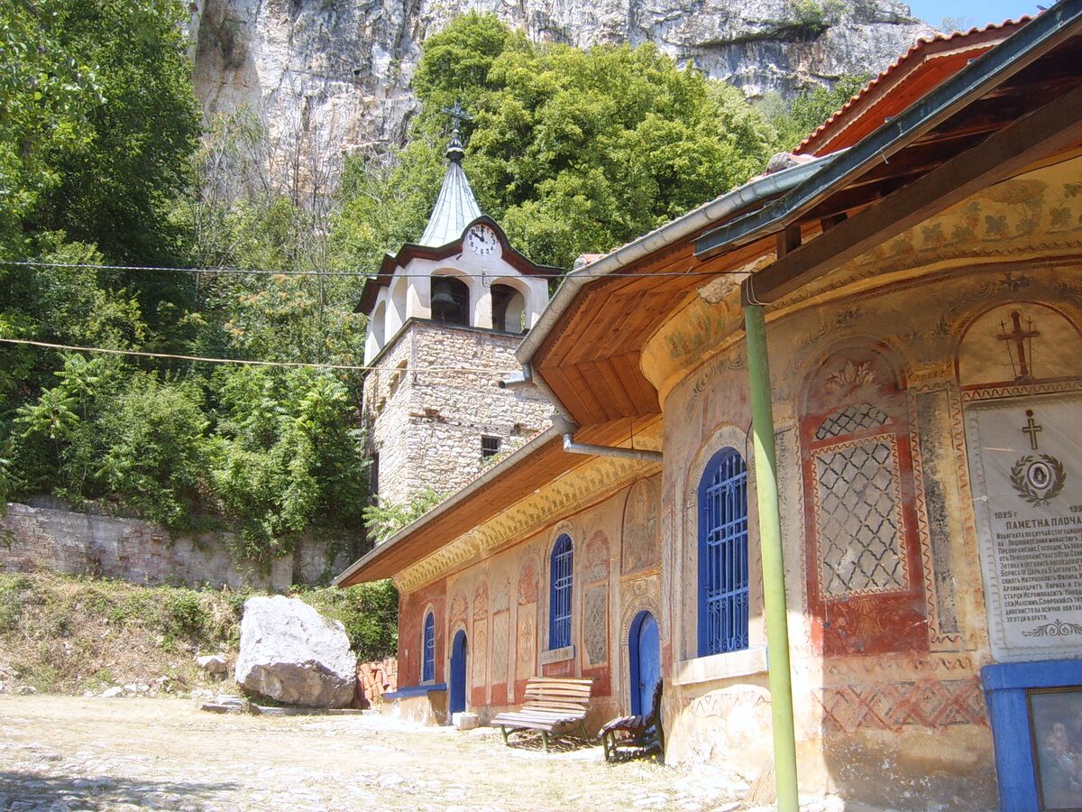 The painted outer walls of the Transfiguration Monastery church and its stone clock tower, set against a sheer limestone cliff above the Yantra gorge.