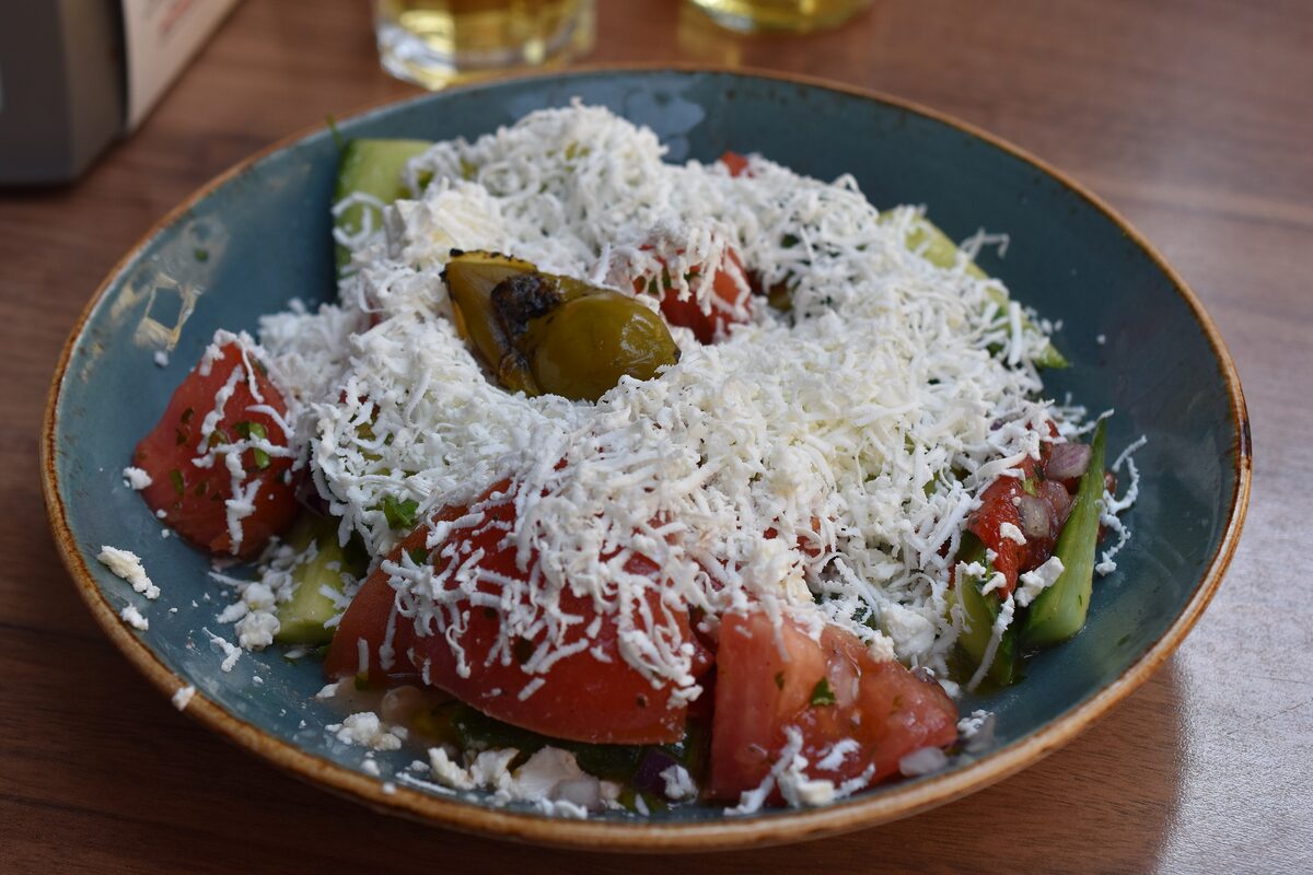 A shopska salad of tomato and cucumber wedges buried under finely grated white sirene cheese, topped with a roasted pepper, on a blue plate.