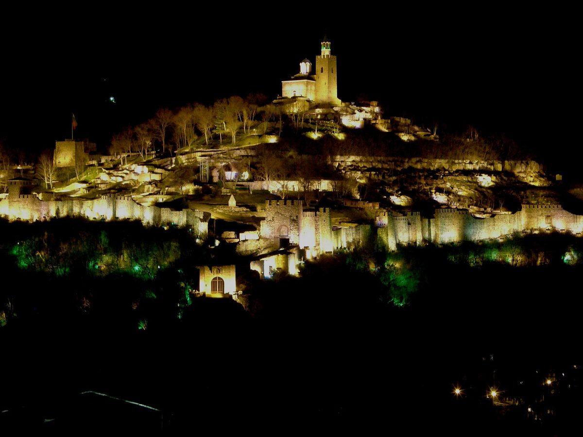 Tsarevets hill lit up at night during the Sound and Light show, the walls glowing gold under the floodlit Patriarchal Church, with green light in the trees below.