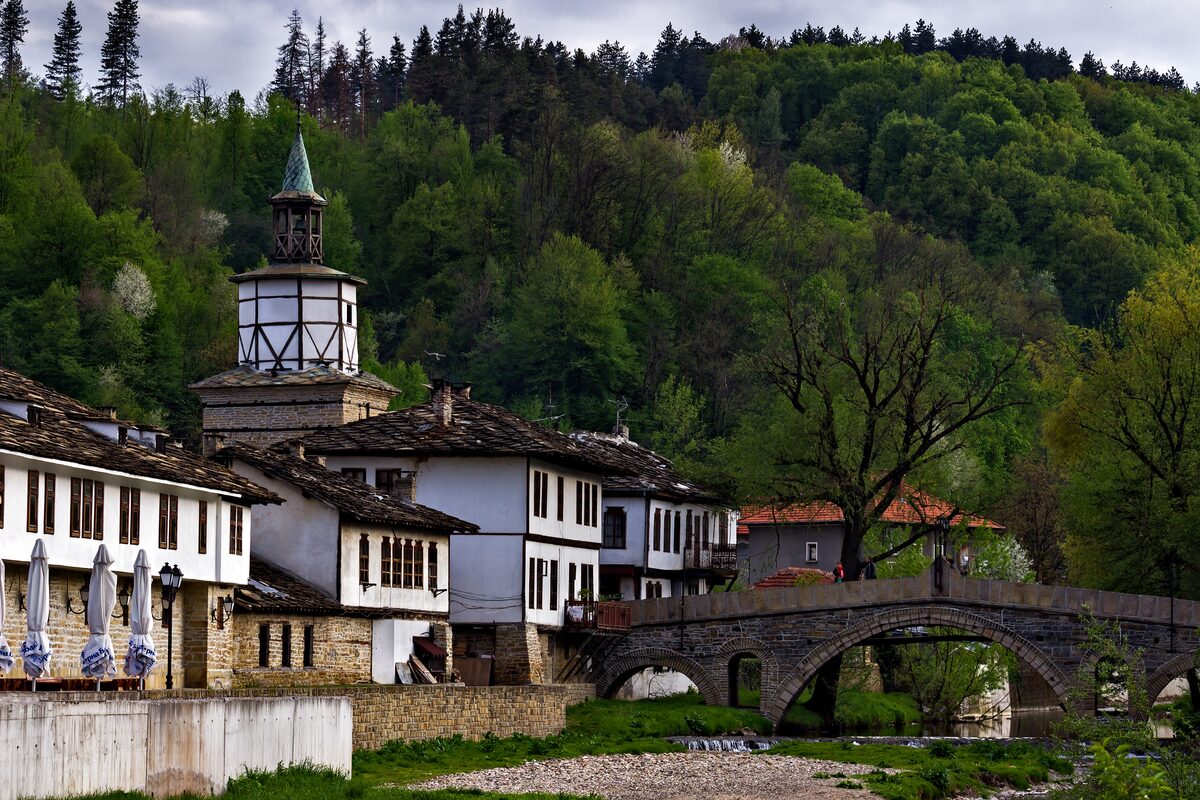 Tryavna's timber-topped clock tower above whitewashed Revival-era houses with stone-slab roofs, beside the arched stone bridge over the river and a forested hill.