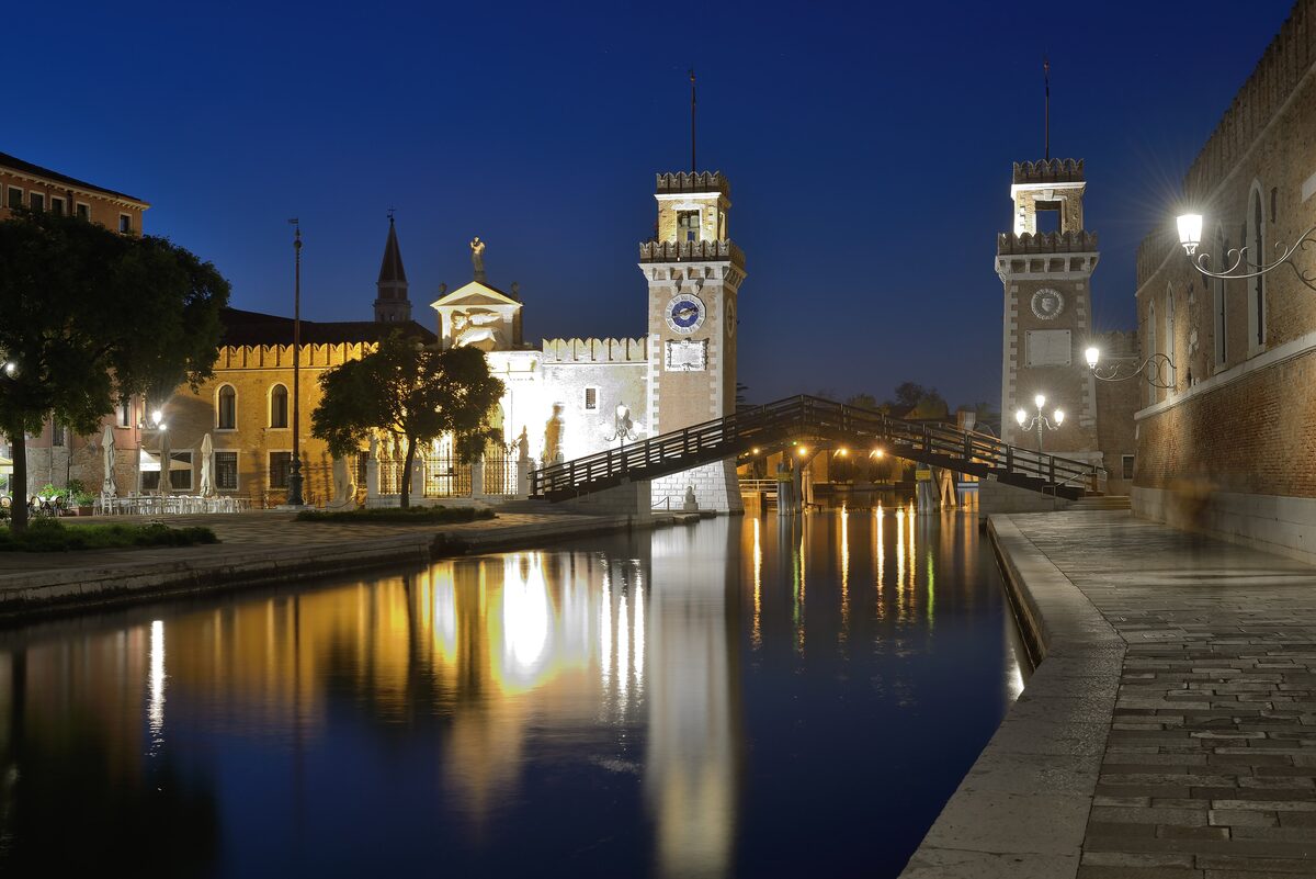 The gateway of the Venetian Arsenale lit up at night between two brick towers, with a wooden bridge over the canal and reflections in the water