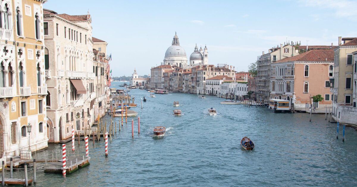 The Grand Canal in Venice on a sunny day, lined with old palaces and striped mooring poles, with motorboats on the water and the domes of Santa Maria della Salute in the distance